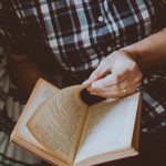 Adult male wearing plaid shirt reading a book indoors for leisure and relaxation.